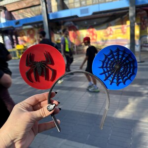 May include: A headband featuring two circular decorations. One is red with a black glitter spider, the other is blue with a black spiderweb design. The headband is silver and held by a hand with black nail polish.