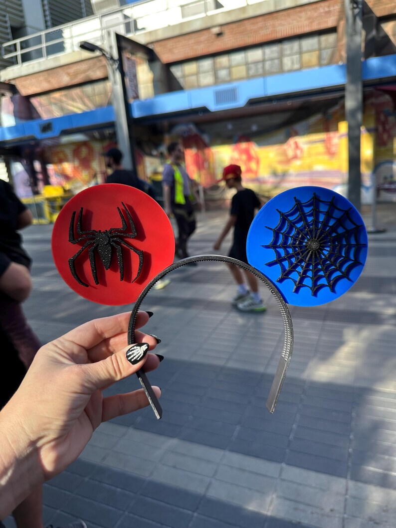 May include: A headband featuring two circular decorations. One is red with a black glitter spider, the other is blue with a black spiderweb design. The headband is silver and held by a hand with black nail polish.