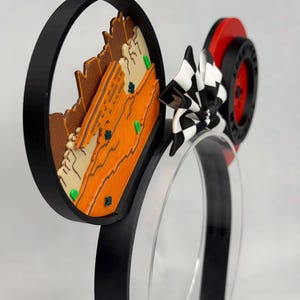 May include: A headband featuring two circular ears. The left ear displays a layered orange and tan desert landscape with small green accents. The right ear features a red wheel with black tire detailing. A black and white checkered bow sits between the ears.