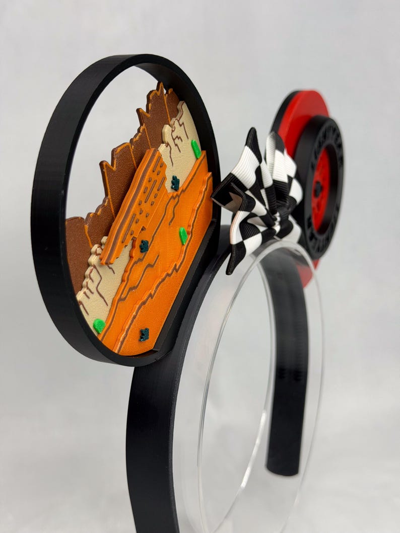 May include: A headband featuring two circular ears. The left ear displays a layered orange and tan desert landscape with small green accents. The right ear features a red wheel with black tire detailing. A black and white checkered bow sits between the ears.