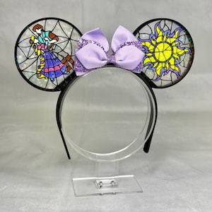Puede incluir: Una diadema con dos orejas circulares con efecto de vidriera. La oreja izquierda muestra a una pareja bailando, mientras que la derecha presenta un emblema de sol amarillo. Un lazo de cinta lavanda con brillos se sit&uacute;a entre las orejas.