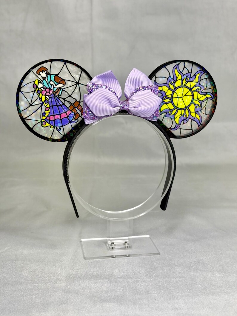 Puede incluir: Una diadema con dos orejas circulares con efecto de vidriera. La oreja izquierda muestra a una pareja bailando, mientras que la derecha presenta un emblema de sol amarillo. Un lazo de cinta lavanda con brillos se sit&uacute;a entre las orejas.