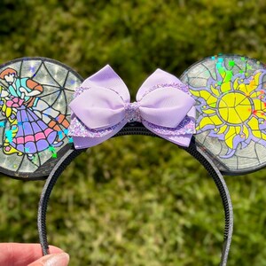 Puede incluir: Una diadema con dos orejas circulares con efecto de vidriera. La oreja izquierda muestra a una pareja bailando, mientras que la derecha exhibe un emblema de sol amarillo. Un lazo morado claro con detalles brillantes se sit&uacute;a entre las orejas.