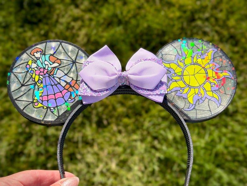 Puede incluir: Una diadema con dos orejas circulares con efecto de vidriera. La oreja izquierda muestra a una pareja bailando, mientras que la derecha exhibe un emblema de sol amarillo. Un lazo morado claro con detalles brillantes se sit&uacute;a entre las orejas.