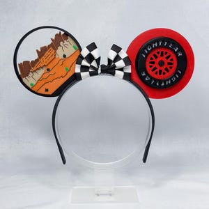 May include: A headband featuring two circular ears and a central black and white checkered bow. The left ear displays a desert landscape with orange rock formations. The right ear features a red and black tire design with the text LIGHTYEAR.