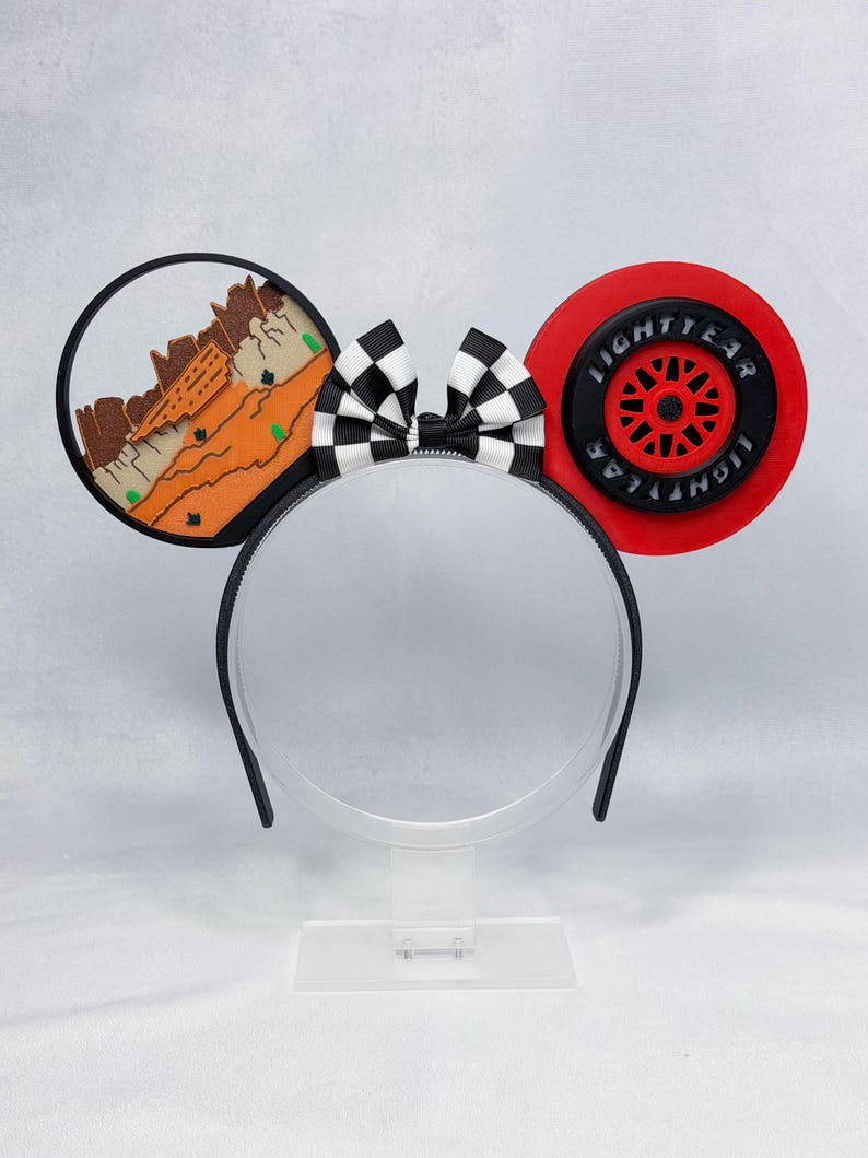 May include: A headband featuring two circular ears and a central black and white checkered bow. The left ear displays a desert landscape with orange rock formations. The right ear features a red and black tire design with the text LIGHTYEAR.