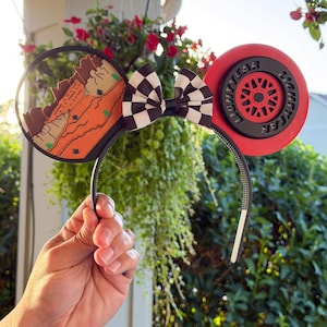 May include: A headband featuring two circular ears. The left ear displays a desert landscape with orange rock formations. The right ear resembles a red racing tire with the text LIGHTYEAR. A black and white checkered bow sits in the center.