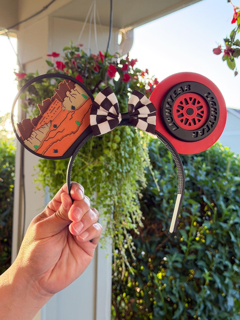 May include: A headband featuring two circular ears. The left ear displays a desert landscape with orange rock formations. The right ear resembles a red racing tire with the text LIGHTYEAR. A black and white checkered bow sits in the center.