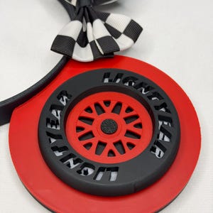 May include: A headband featuring a black and white checkered bow and a circular red base. The center of the base displays a black tire with the word LIGHTYEAR embossed on it, surrounding a red wheel with multiple spokes and a black center hub.