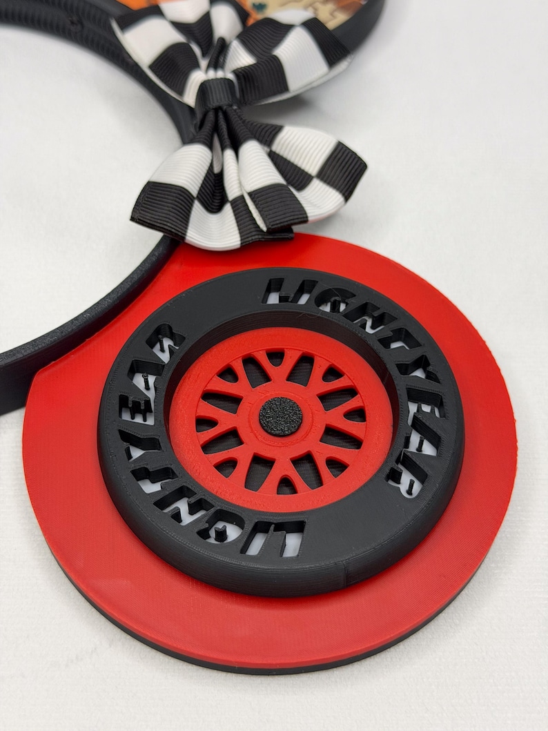 May include: A headband featuring a black and white checkered bow and a circular red base. The center of the base displays a black tire with the word LIGHTYEAR embossed on it, surrounding a red wheel with multiple spokes and a black center hub.
