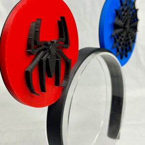 May include: A pair of black and red Spider-Man themed mouse ears. The ears are circular, with a red ear featuring a black spider emblem and a blue ear with a black spiderweb design. The headband is black.