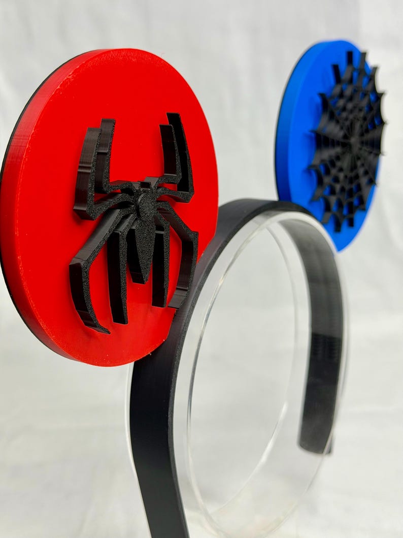 May include: A pair of black and red Spider-Man themed mouse ears. The ears are circular, with a red ear featuring a black spider emblem and a blue ear with a black spiderweb design. The headband is black.