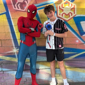 May include: A person in a Spider-Man costume stands next to a person wearing a baseball jersey with a Spider-Man graphic. The background features colorful graffiti art with Spider-Man-themed designs. The person in the jersey is holding a Spider-Man fan.