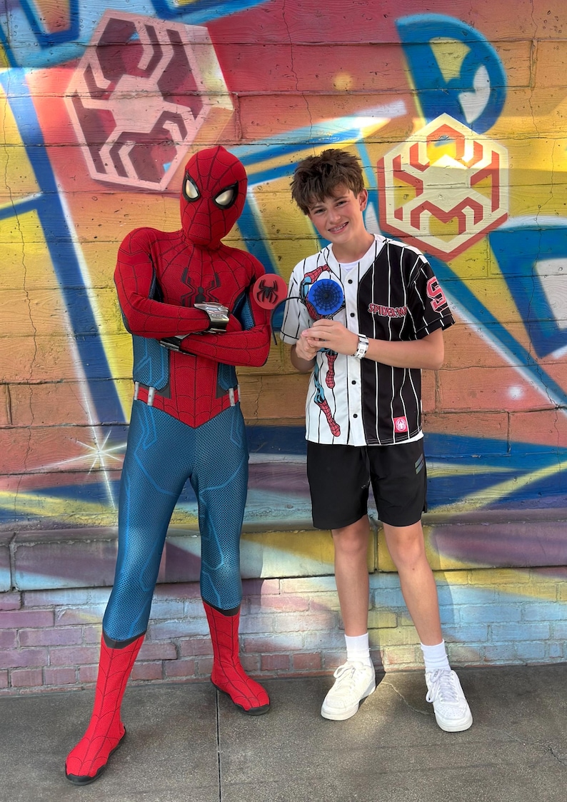 May include: A person in a Spider-Man costume stands next to a person wearing a baseball jersey with a Spider-Man graphic. The background features colorful graffiti art with Spider-Man-themed designs. The person in the jersey is holding a Spider-Man fan.