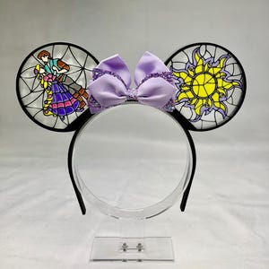 Puede incluir: Una diadema con dos orejas circulares con efecto de vidriera. La oreja izquierda muestra a una pareja bailando, mientras que la derecha exhibe un emblema de sol amarillo. Un lazo de tela lavanda con detalles morados brillantes se sit&uacute;a entre las orejas.