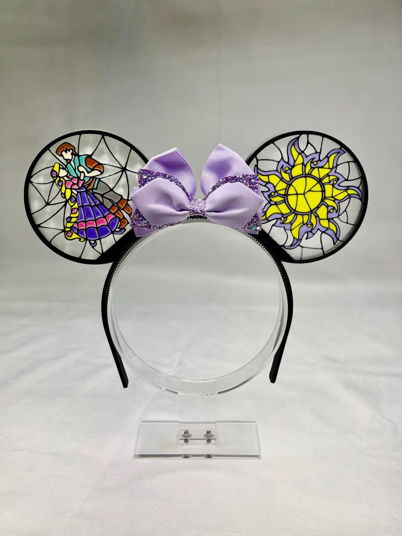Puede incluir: Una diadema con dos orejas circulares con efecto de vidriera. La oreja izquierda muestra a una pareja bailando, mientras que la derecha exhibe un emblema de sol amarillo. Un lazo de tela lavanda con detalles morados brillantes se sit&uacute;a entre las orejas.