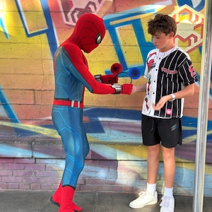 May include: A person dressed as Spider-Man is offering a pair of Spider-Man-themed ear headbands to a young person. The background features a colorful graffiti wall. The Spider-Man costume is red and blue.