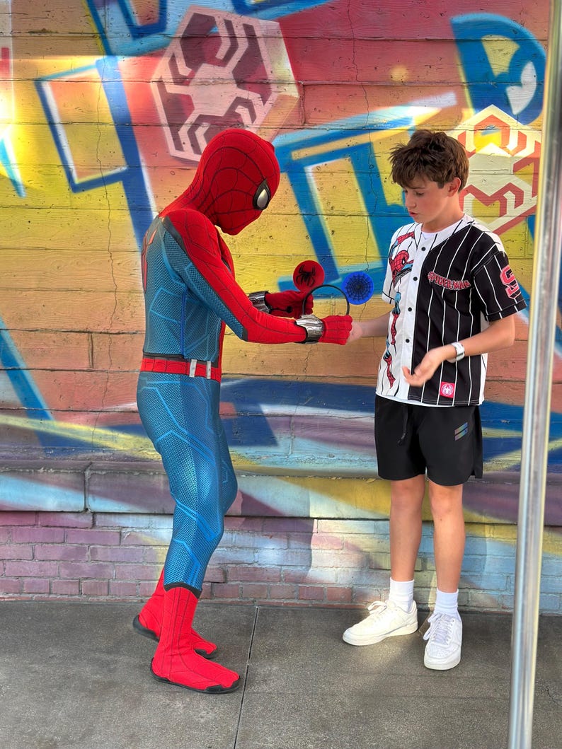 May include: A person dressed as Spider-Man is offering a pair of Spider-Man-themed ear headbands to a young person. The background features a colorful graffiti wall. The Spider-Man costume is red and blue.
