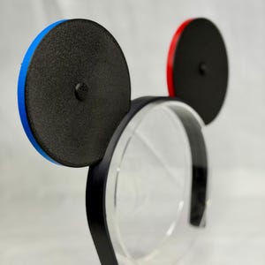 May include: Black mouse ear headband with one blue and one red rim. The ears are black with a textured surface. The headband is black and sits on a clear display stand.