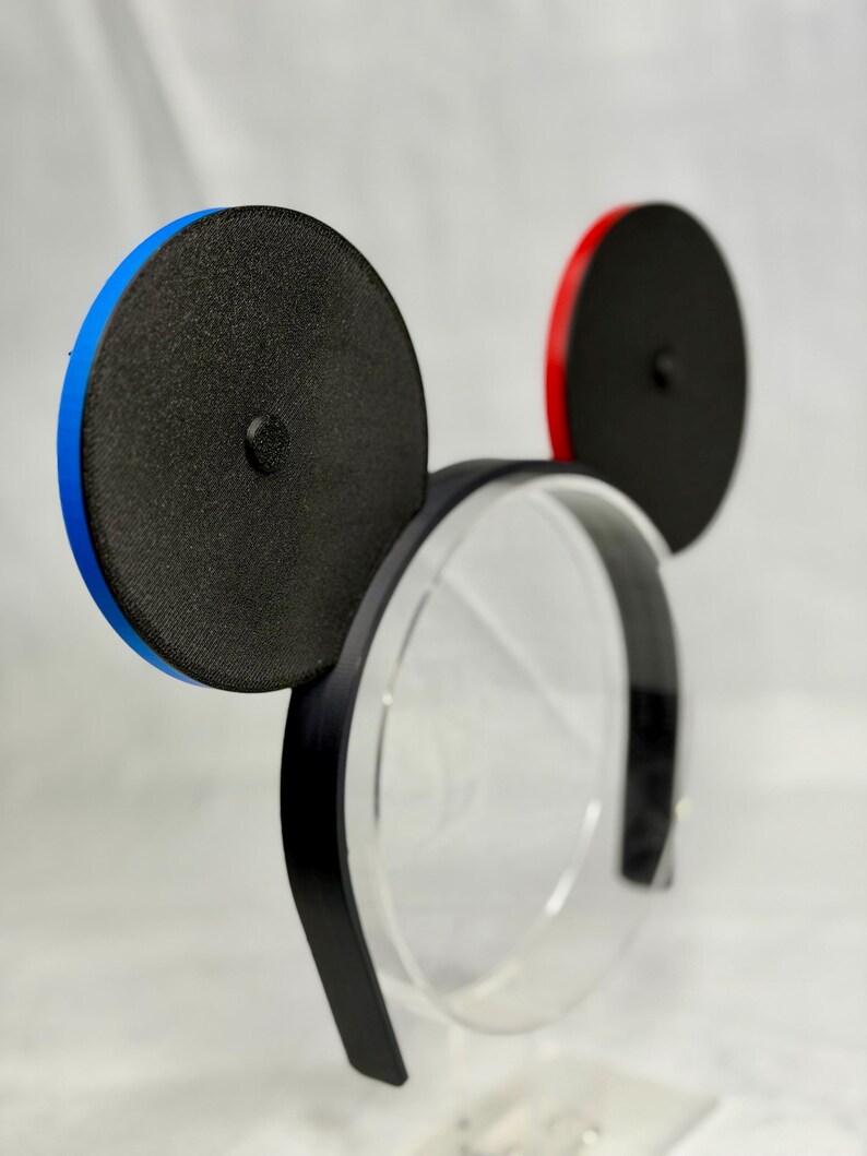 May include: Black mouse ear headband with one blue and one red rim. The ears are black with a textured surface. The headband is black and sits on a clear display stand.