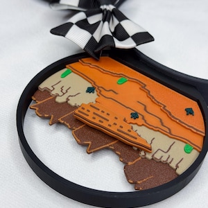 May include: A black headband featuring circular ears with a desert landscape design in shades of orange, tan, and brown. The ears are accented with a black and white checkered bow. The landscape includes small green cacti and a central building structure.