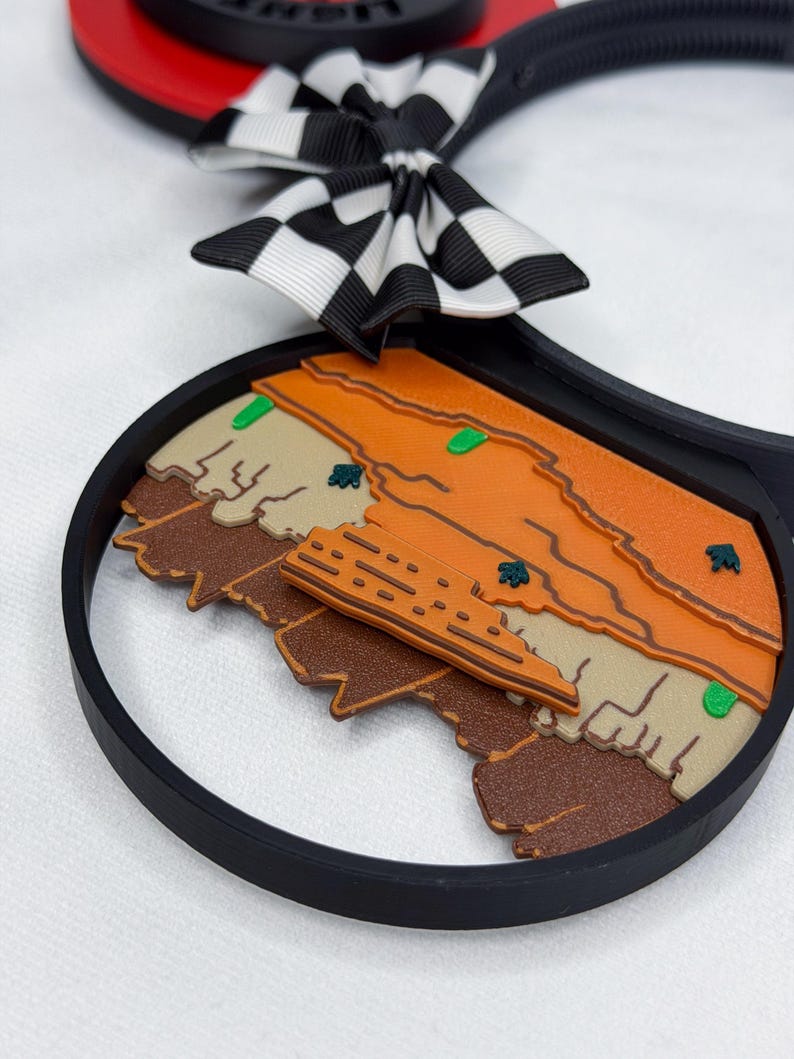 May include: A black headband featuring circular ears with a desert landscape design in shades of orange, tan, and brown. The ears are accented with a black and white checkered bow. The landscape includes small green cacti and a central building structure.