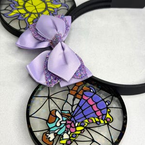 Puede incluir: Una diadema con dos orejas circulares con efecto de vidriera. Una oreja muestra un dise&ntilde;o de sol amarillo, mientras que la otra muestra un personaje con vestido morado. Un lazo de cinta lavanda con detalles brillantes se sit&uacute;a entre las orejas.