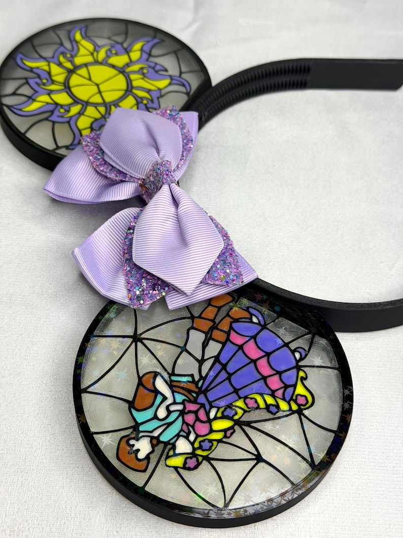 Puede incluir: Una diadema con dos orejas circulares con efecto de vidriera. Una oreja muestra un dise&ntilde;o de sol amarillo, mientras que la otra muestra un personaje con vestido morado. Un lazo de cinta lavanda con detalles brillantes se sit&uacute;a entre las orejas.