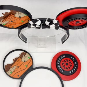 May include: A headband with two circular ears featuring interchangeable designs. One ear displays a desert landscape, the other a red and black wheel with the text LIGHTYEAR. A black and white checkered bow sits in the center. Two extra ear attachments are included.