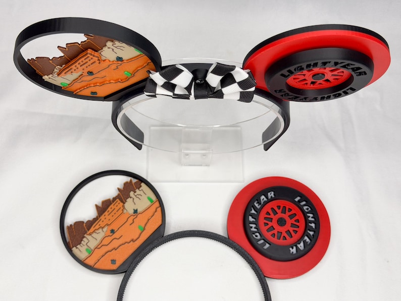 May include: A headband with two circular ears featuring interchangeable designs. One ear displays a desert landscape, the other a red and black wheel with the text LIGHTYEAR. A black and white checkered bow sits in the center. Two extra ear attachments are included.