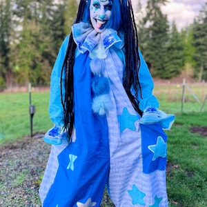 May include: A blue and white clown costume with star accents. The costume features a light blue top with a ruffled collar and a matching jumpsuit with star-shaped patches. The clown has blue hair and face paint.