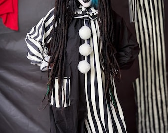 May include: A person in a black and white striped clown costume with dreadlocks and white pom-poms. The costume features a black and white striped top and pants, with a black collar and white pom-poms down the front. The person has white face paint with black accents.