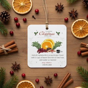 Peut inclure: Ornement de Noël blanc avec le texte "Stovetop Christmas Potpourri" et des instructions. L'ornement présente des illustrations de tranches d'orange, de canneberges et de bâtons de cannelle, entourées de décorations festives.