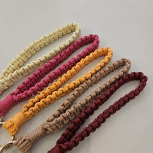Pu&ograve; includere: Primo piano di sei cordini da polso in macram&egrave; di vari colori. I colori includono beige, magenta, arancione, marrone chiaro e bordeaux. Ogni cordino &egrave; realizzato con cordoncino annodato e ha un anello per il polso. I cordini sono un artigianato fatto a mano.