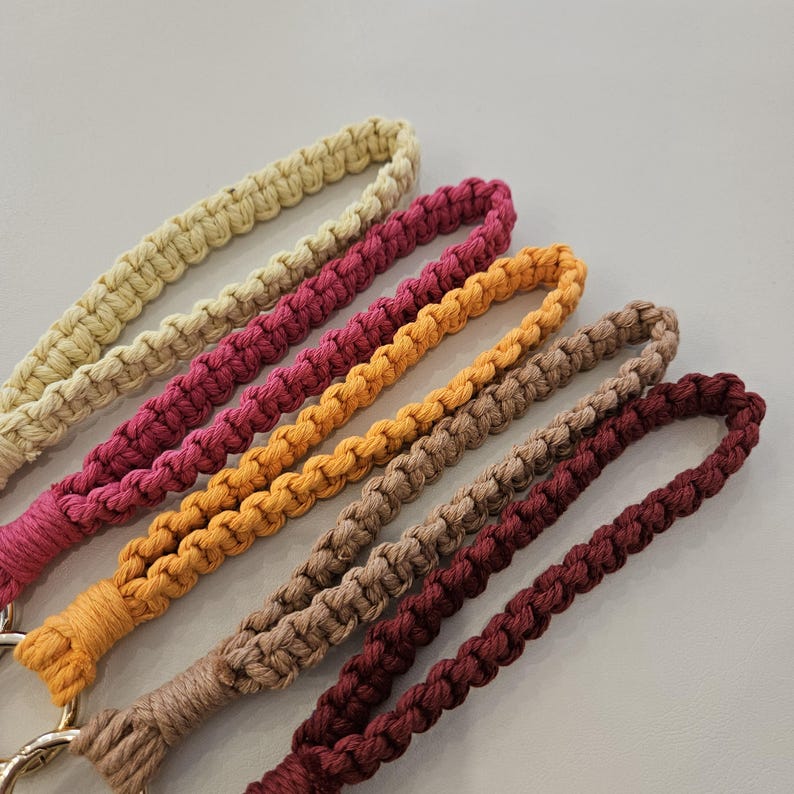 Pu&ograve; includere: Primo piano di sei cordini da polso in macram&egrave; di vari colori. I colori includono beige, magenta, arancione, marrone chiaro e bordeaux. Ogni cordino &egrave; realizzato con cordoncino annodato e ha un anello per il polso. I cordini sono un artigianato fatto a mano.