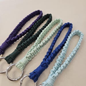 Pu&ograve; includere: Primo piano di cinque cordini macram&egrave; in vari colori: viola, verde scuro, verde chiaro, blu e azzurro. Ogni cordino ha un anello metallico color argento alla base. Il macram&egrave; &egrave; intrecciato fittamente, creando un aspetto strutturato.