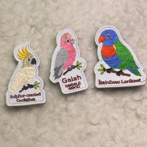 May include: Three embroidered patches featuring birds on branches. The left patch shows a Sulphur-crested Cockatoo, the centre patch shows a pink and grey Galah, and the right patch shows a colourful Rainbow Lorikeet with blue, orange, and green feathers.