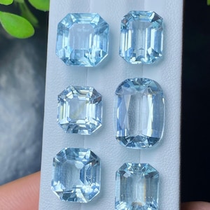 Puede incluir: Seis piedras preciosas de aguamarina azul claro, con diferentes cortes rectangulares y esmeralda, se muestran sobre una superficie blanca. Las gemas están facetadas, mostrando su claridad y brillo. Las piedras preciosas están dispuestas en dos filas de tres.