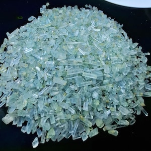 Peut inclure: Un tas de petites pierres précieuses cristallines bleu-vert clair. Les cristaux sont rectangulaires et semblent bruts, de tailles et de formes variées. Les pierres précieuses sont dispersées sur une surface sombre, créant un contraste.
