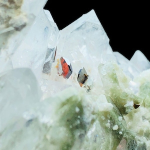 Puede incluir: Primer plano de un grupo de cristales de cuarzo con formaciones claras y puntiagudas. El cristal tiene inclusiones de minerales rojos y marrones, sobre un fondo negro. Se ven depósitos minerales verdes y blancos.