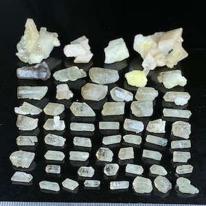 Könnte beinhalten: Eine Sammlung roher, ungeschliffener Edelsteine in verschiedenen Formen und Größen. Die Steine reichen von klar bis hellgelb und sind auf einer schwarzen Oberfläche angeordnet. Ein Lineal am Boden dient als Maßstab.