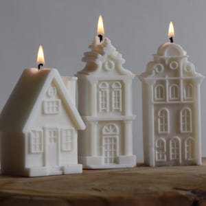 Puede incluir: Tres velas blancas con forma de casa con mechas encendidas. Las velas son de diferentes tamaños y estilos arquitectónicos, parecidas a edificios en miniatura. Se colocan sobre una superficie de madera sobre un fondo neutro.