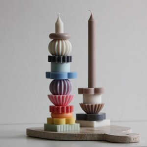 Puede incluir: Una colorida composición de velas con dos candelabros. Uno es una torre apilada de varias formas y colores, incluyendo blanco, negro, azul, morado, rosa, naranja y amarillo. El otro es una vela marrón con una base en forma de flor.
