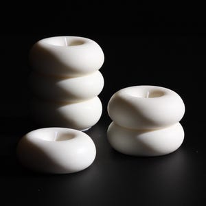 Könnte beinhalten: Drei weiße Donut-förmige Kerzen, zwei übereinander gestapelt und eine einzelne Kerze. Die Kerzen stehen auf einer schwarzen Oberfläche.