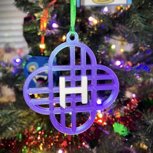 May include: A purple and white Christmas ornament in the shape of a four-leaf clover with the letter "H" in the center. The ornament is hanging on a Christmas tree with a green ribbon.