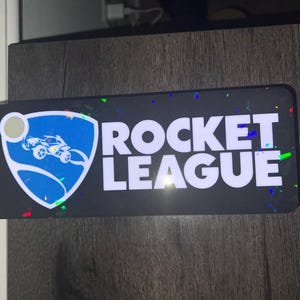 May include: A rectangular black sticker with the words "ROCKET LEAGUE" in large white letters. The sticker also features the Rocket League logo, a blue shield with a white car and a circle. Colorful confetti-like accents are scattered around the edges.