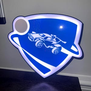 Puede incluir: Un letrero iluminado azul y blanco con un coche estilizado dentro de un escudo. El letrero tiene un borde blanco y un elemento circular en la parte superior. El diseño recuerda a un logotipo de videojuego.