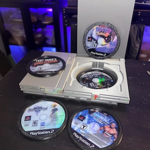 May include: A silver PlayStation 2 console with five game discs. The games include "Spyro," "Battlefront," "Tony Hawk's Underground," "Kingdom Hearts," and "SmackDown vs. Raw." The discs are in black holders.