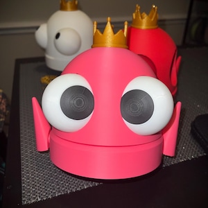 May include: A pink, white, and red cartoon character with large white eyes and a gold crown. The character has a round body and two small side fins. The eyes have black pupils. The character is sitting on a dark surface.