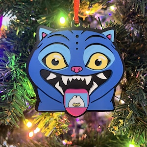 May include: A blue cat-shaped Christmas ornament with a cartoonish design. The ornament has large yellow eyes, sharp white teeth, and a pink tongue with a small image. It hangs from a red ribbon, perfect for holiday decor.